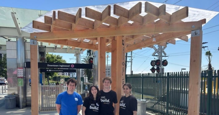 Four students stand underneath a wooden shade structure at the Downtown-Inglewood Metro station