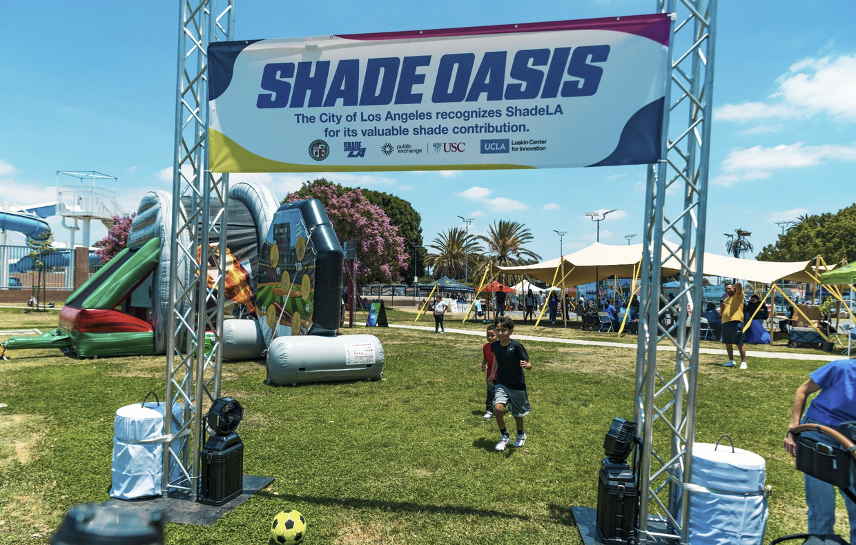 Two kids run under metal arch with sign that reads 'Shade Oasis The City of Los Angeles recognizes ShadeLA for its valuable shade contribution.'