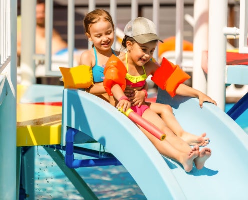 Two children in the sun wearing swimsuits and flotation devices slide down a water slide into a pool.