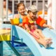 Two children in the sun wearing swimsuits and flotation devices slide down a water slide into a pool.