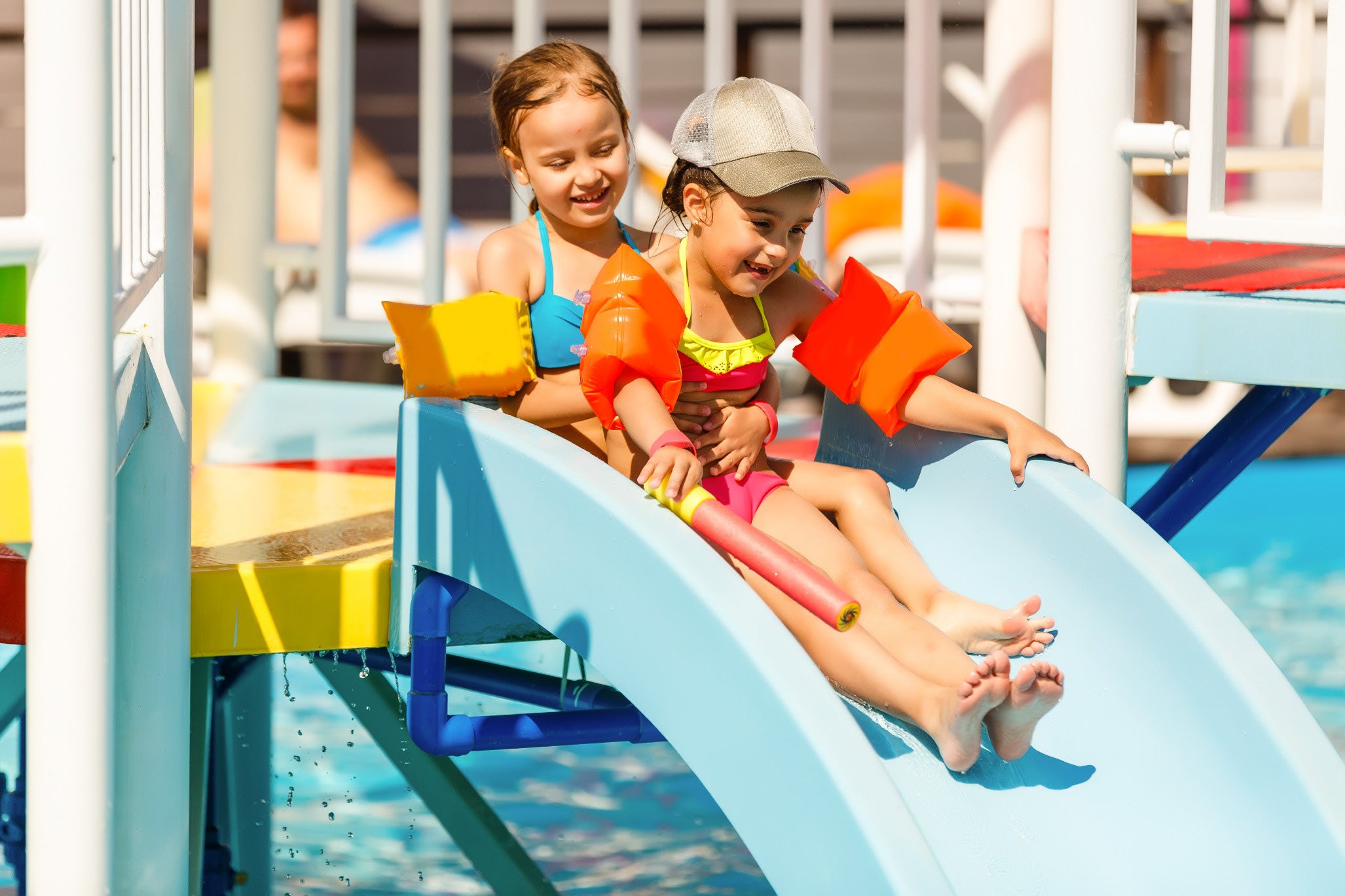 Two children in the sun wearing swimsuits and flotation devices slide down a water slide into a pool.