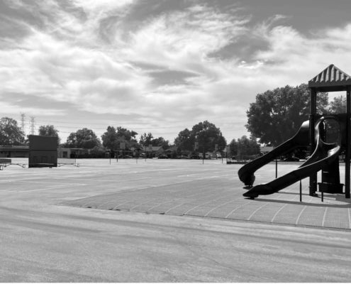 Unshaded playground with two slides and a large paved surface, with trees and buildings in the background.